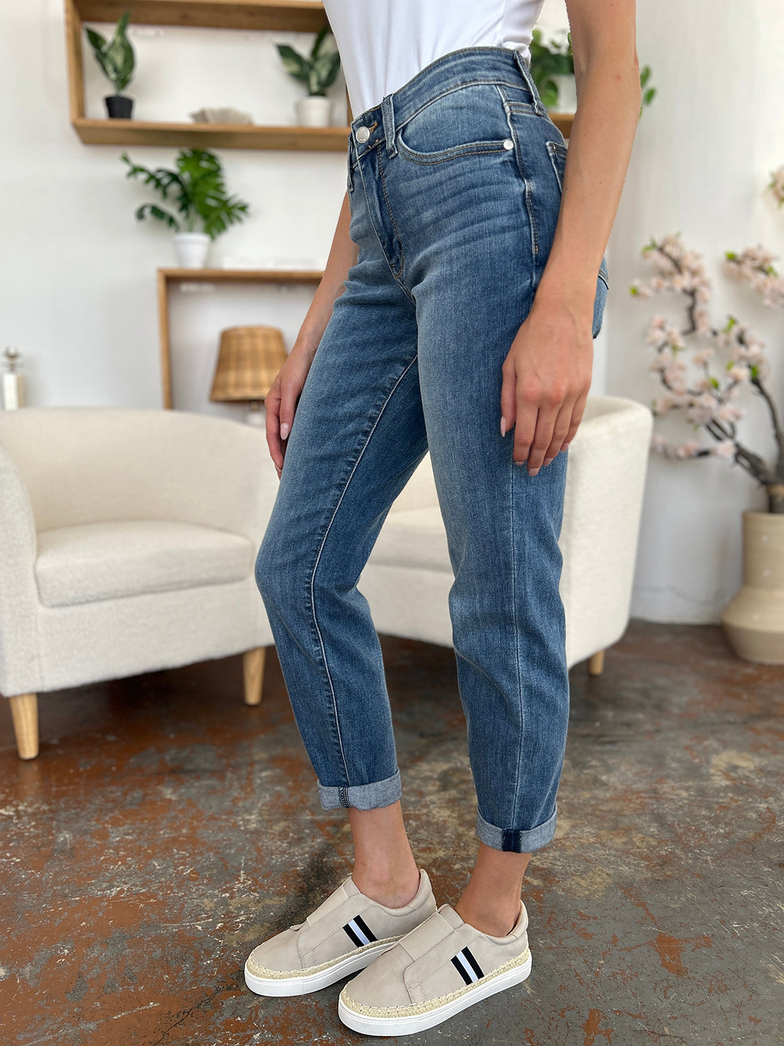 A person wearing a white shirt, Judy Blue Full Size Cuffed Hem Slim Jeans, and black wedge-heeled sandals is standing in a room with beige chairs and plants in the background.