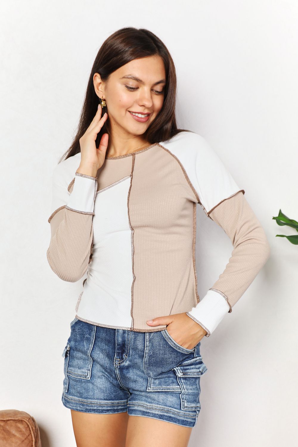 A woman with long brown hair is wearing the "Double Take Color Block Exposed Seam Top" in beige and white, paired with jeans. She stands with one hand in her pocket and the other touching her hair, smiling. A plant is visible in the background.