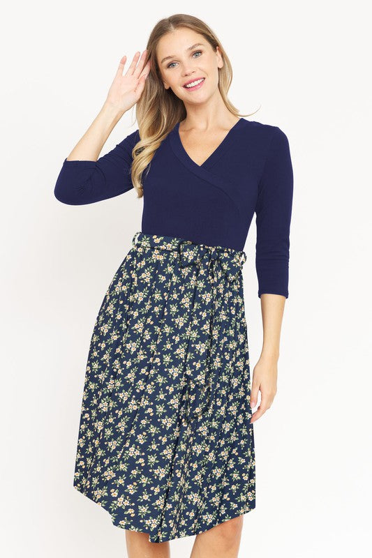 A woman in a Surplice Floral Sash Midi Dress, comprised of 95% Polyester and 5% Spandex, stands against a plain white backdrop. The knee-length dress, designed in navy blue with a floral skirt and wrapped bodice, showcases elegant craftsmanship from the United States.