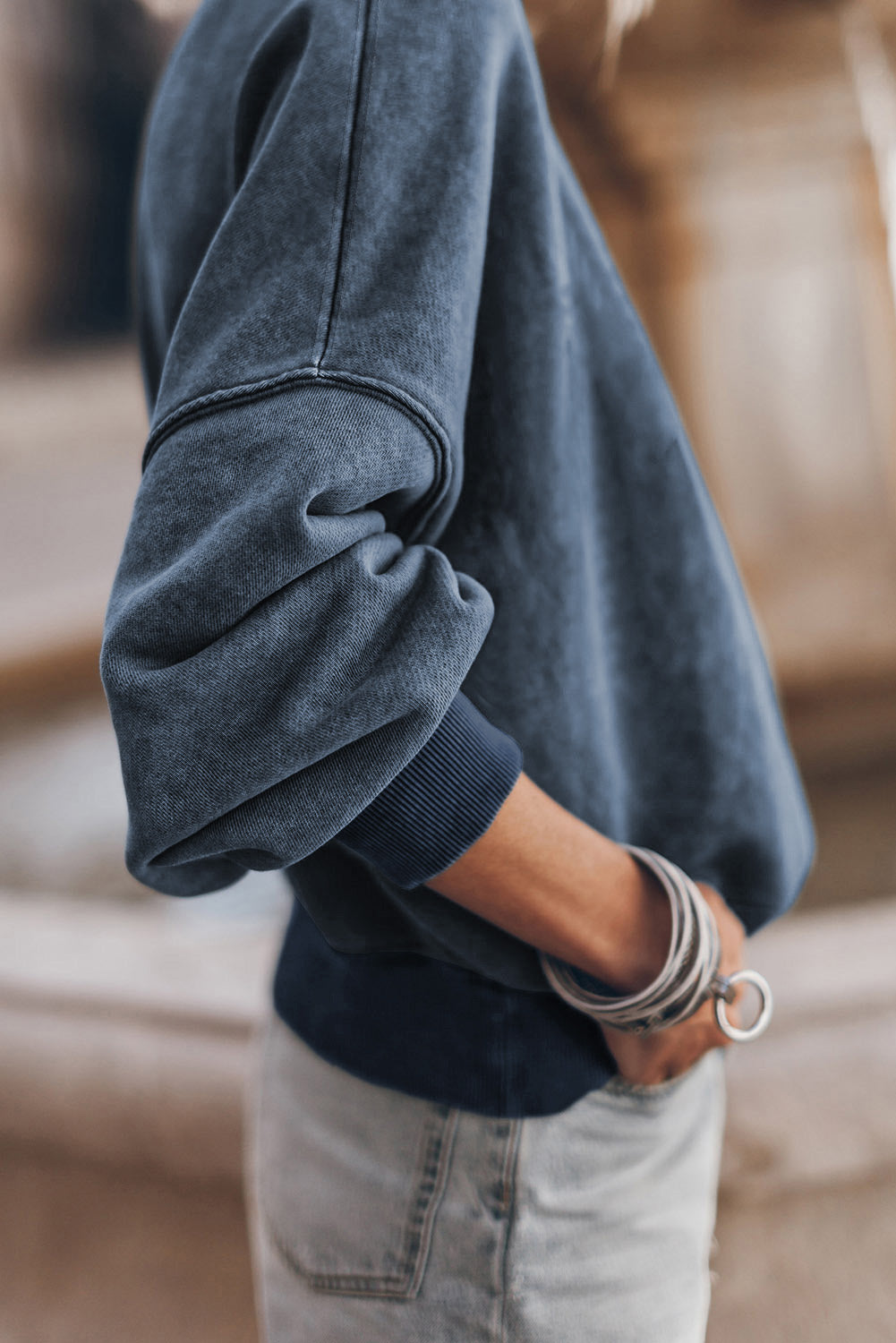 A person with long blonde hair is wearing a sky blue drop shoulder crew neck pullover sweatshirt and light denim shorts, standing outdoors with their back to the camera.