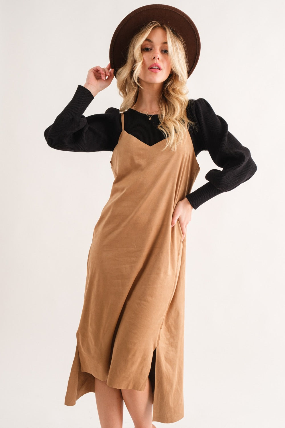 A woman wearing a wide-brimmed hat, black long-sleeve top, and the And The Why Faux Suede Cut Edge Slit Cami Midi Dress poses against a plain background.