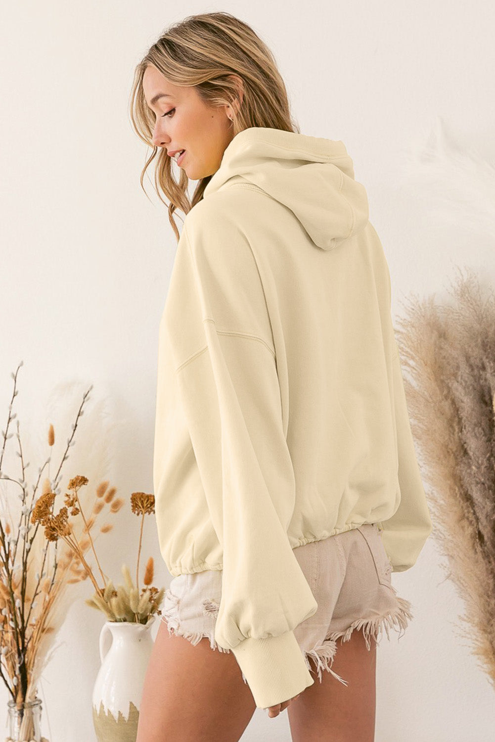 A person wearing a loose fit Beige Ribbed Trim Kangaroo Pocket Zipped Hoodie and frayed shorts stands sideways, with dried plants and a vase in the background.