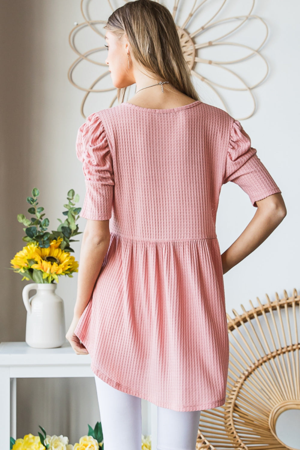 A woman in a Heimish Full Size Waffle Knit V-Neck Babydoll Top with short-sleeves and a long necklace, standing by a window with a neutral expression; the perfect cozy wardrobe addition.