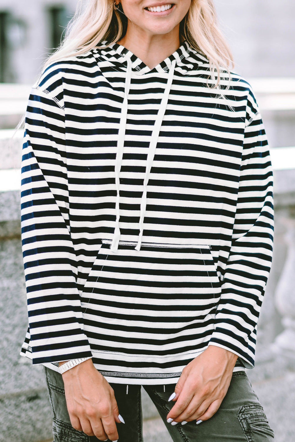 A person with long, blonde hair wearing a trendy Stripe Kangaroo Pocket Drawstring Hoodie with black and white stripes and dark green pants is standing outdoors, facing away from the camera.