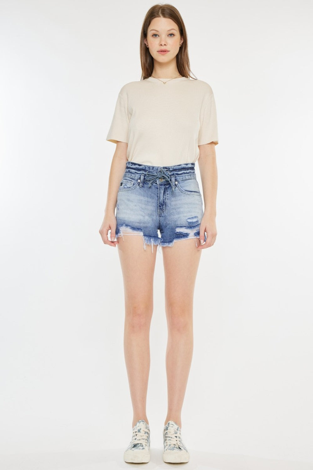 A person wearing a beige t-shirt and Kancan High Rise Frayed Hem Denim Shorts, paired with patterned sneakers, stands against a white background—an ideal addition to any summer wardrobe.