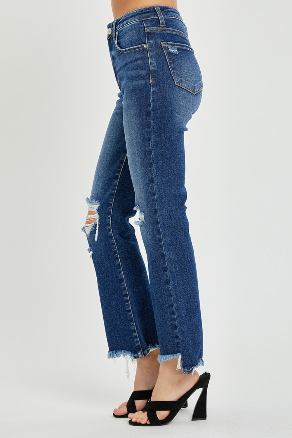 A person wearing RISEN High Rise Distressed Crop Flare Jeans with frayed hems stands with their right hand on their left hip, paired with black open-toed sandals.