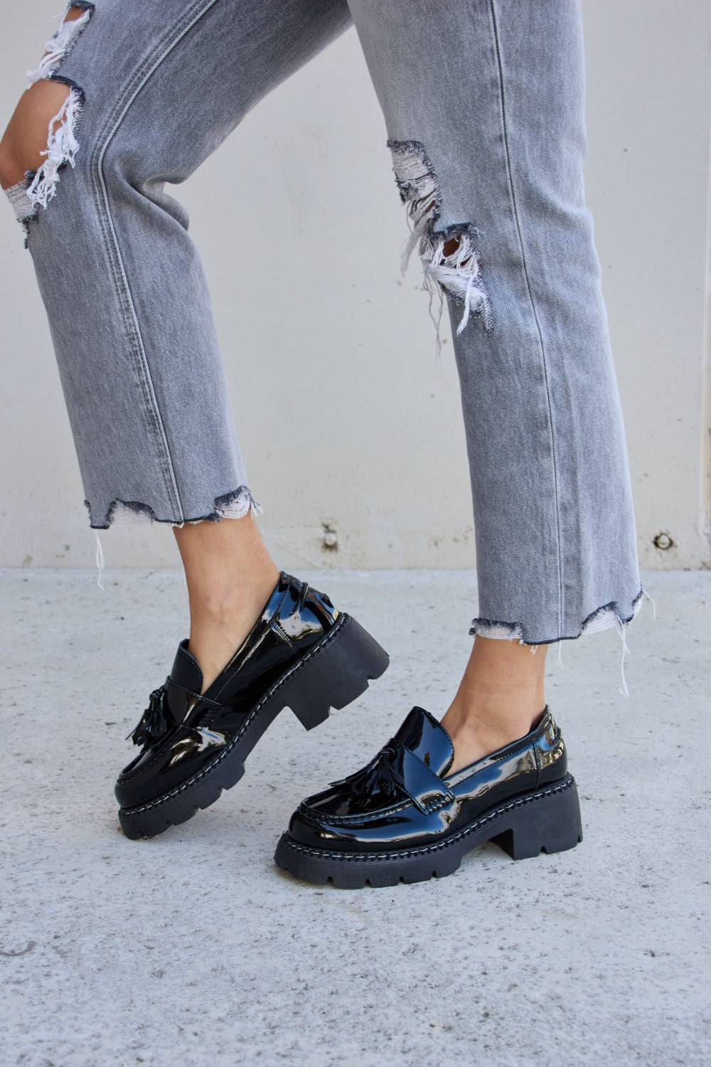 A person wearing ripped gray jeans and the Forever Link Tassel Detail Mid Heel Chunky Loafers, which feature shiny black tones, chunky soles, and a subtle tassel detail.