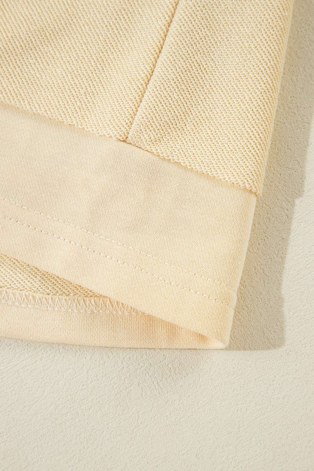 A woman with wavy hair stands with her back to the camera, wearing a Beige Splicing Round Neck Pullover Sweatshirt and blue jeans. She is in front of a white wall and a decorative plant, presenting a casual yet polished look.