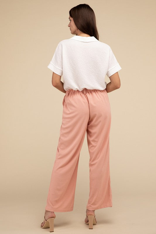 A woman stands against a beige background, highlighting her chic ensemble with a tailored fit. She wears a white short-sleeve shirt paired with versatile pink Waffle Trouser Pants, hands casually in pockets, complemented by stylish strappy sandals.