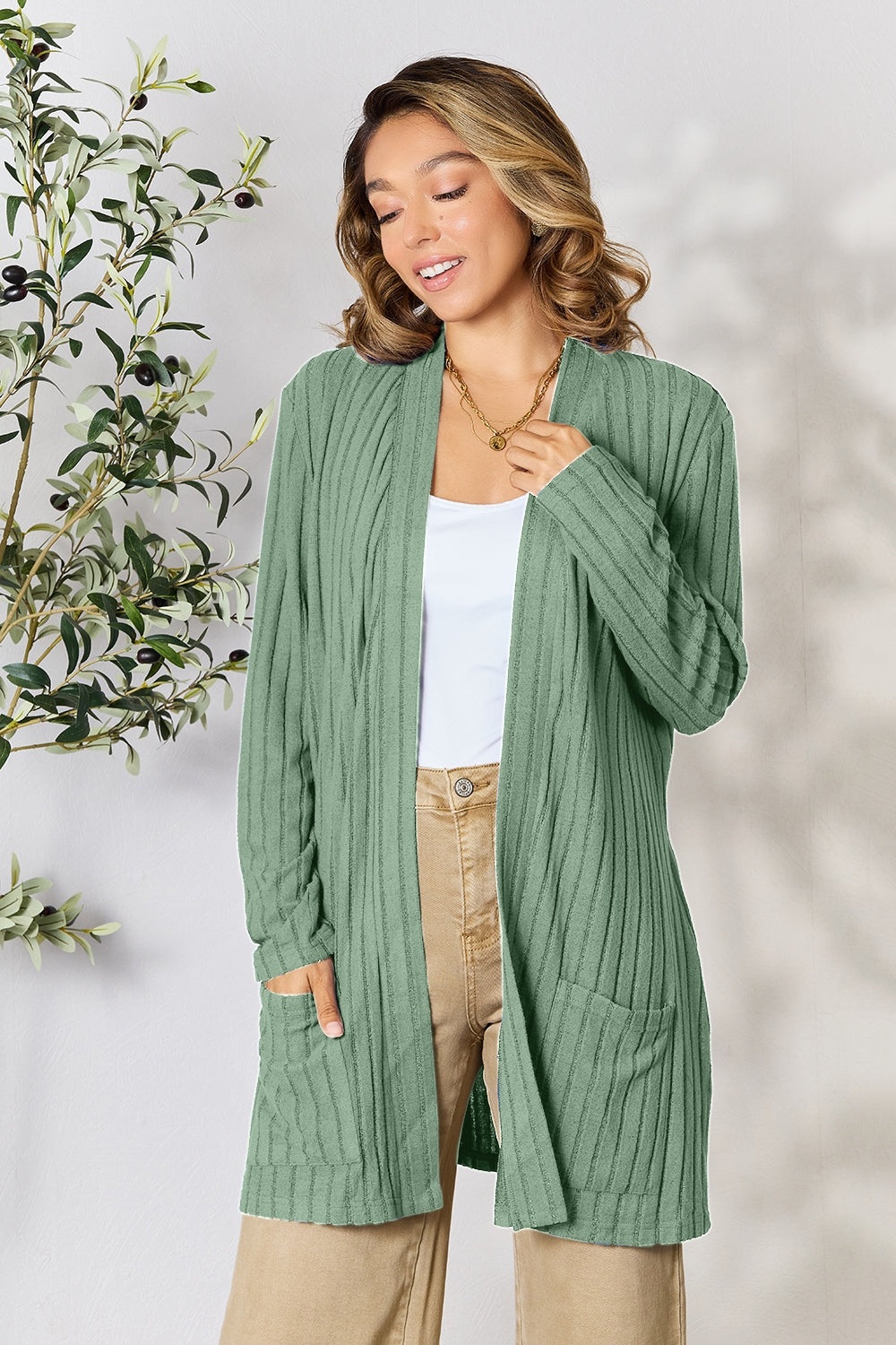 A woman in a Basic Bae Full Size Ribbed Open Front Cardigan with Pockets and jeans, made from stretchy material, stands in front of a plant, smiling slightly and looking to the side.