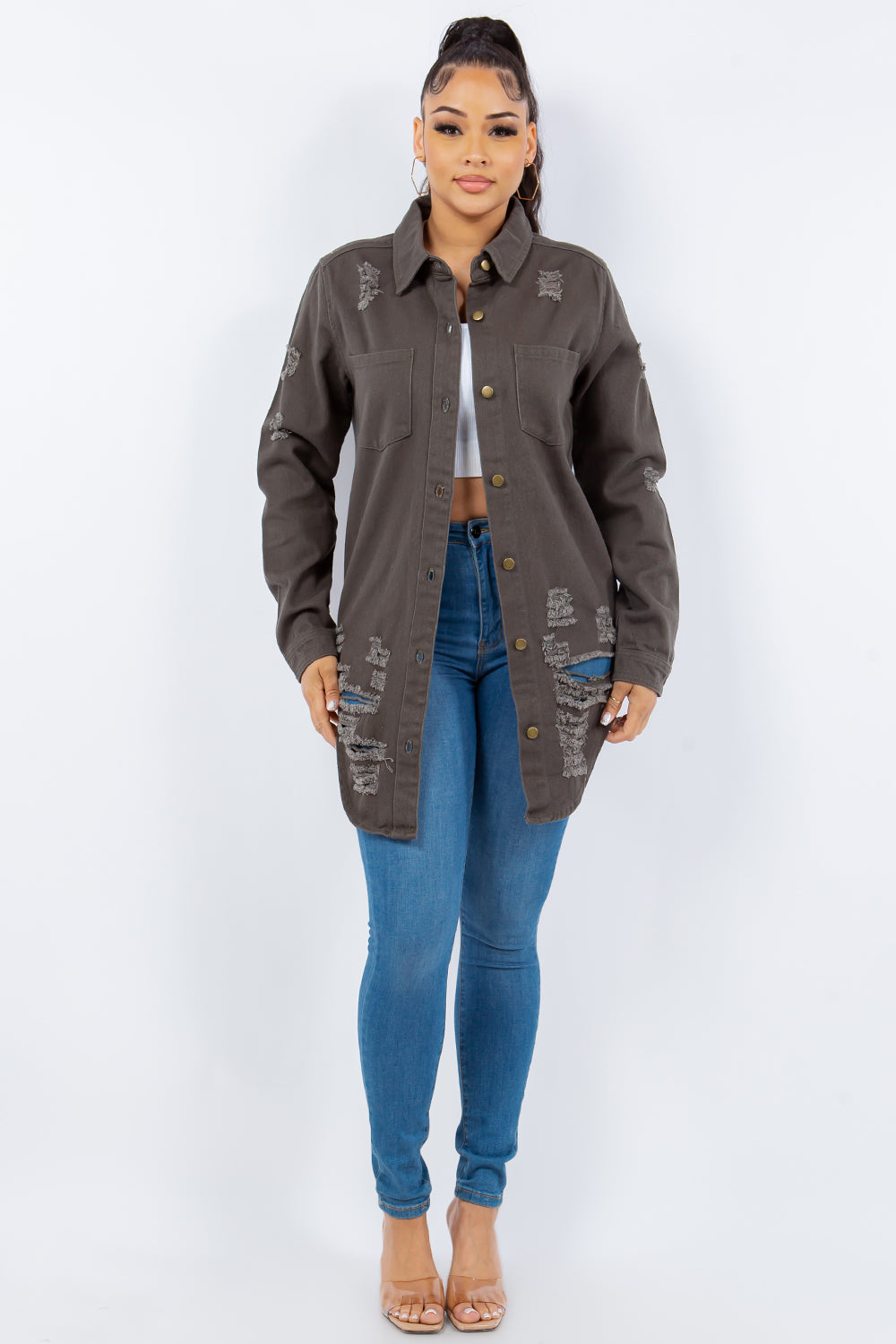 A woman stands against a light background wearing the American Bazi Distressed Button Up Long Sleeve Denim Jacket with a white crop top and blue ripped jeans.