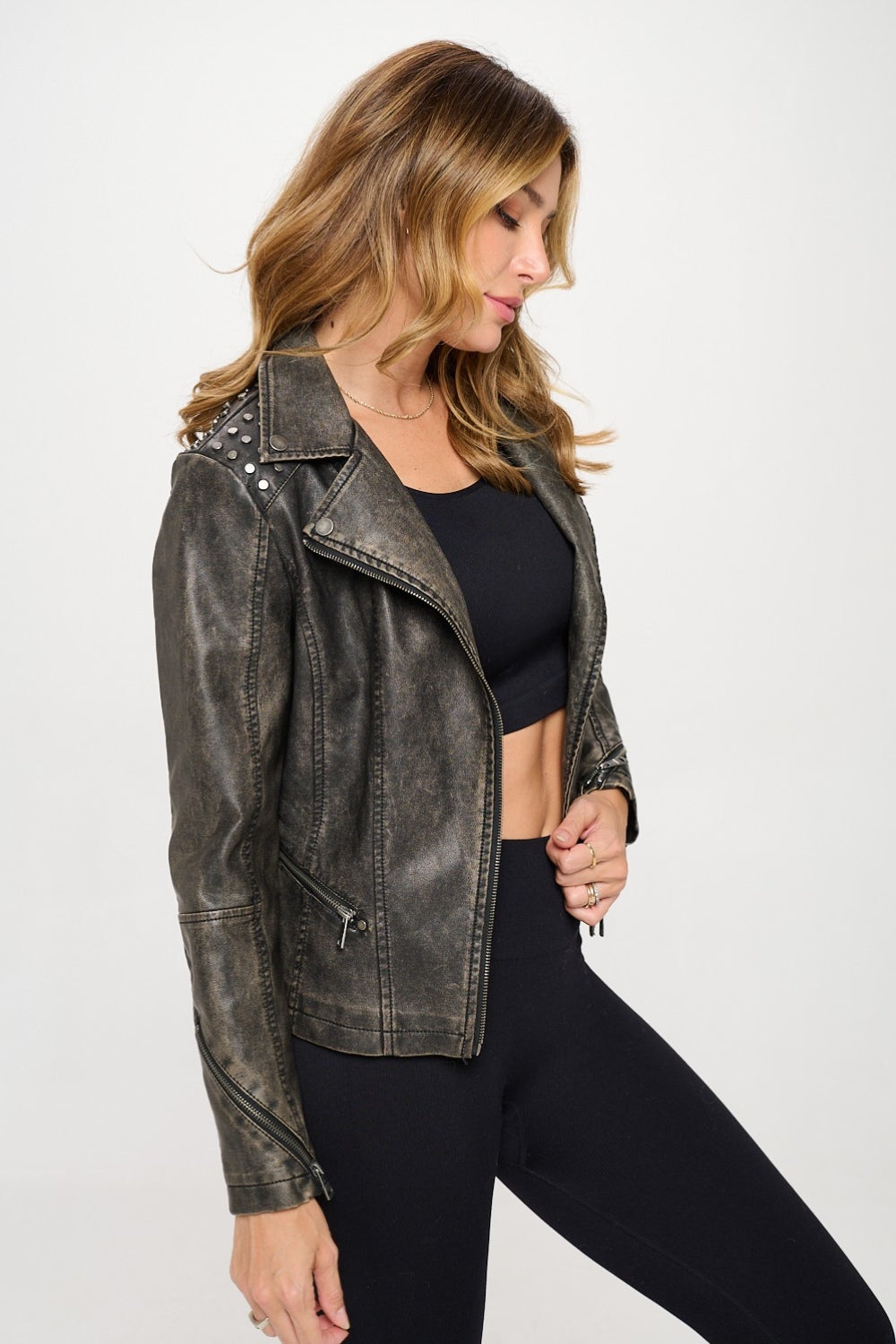 A person wearing the Coalition LA Studded Classic Moto Faux Leather Jacket, along with a black crop top and black pants, stands against a white background.