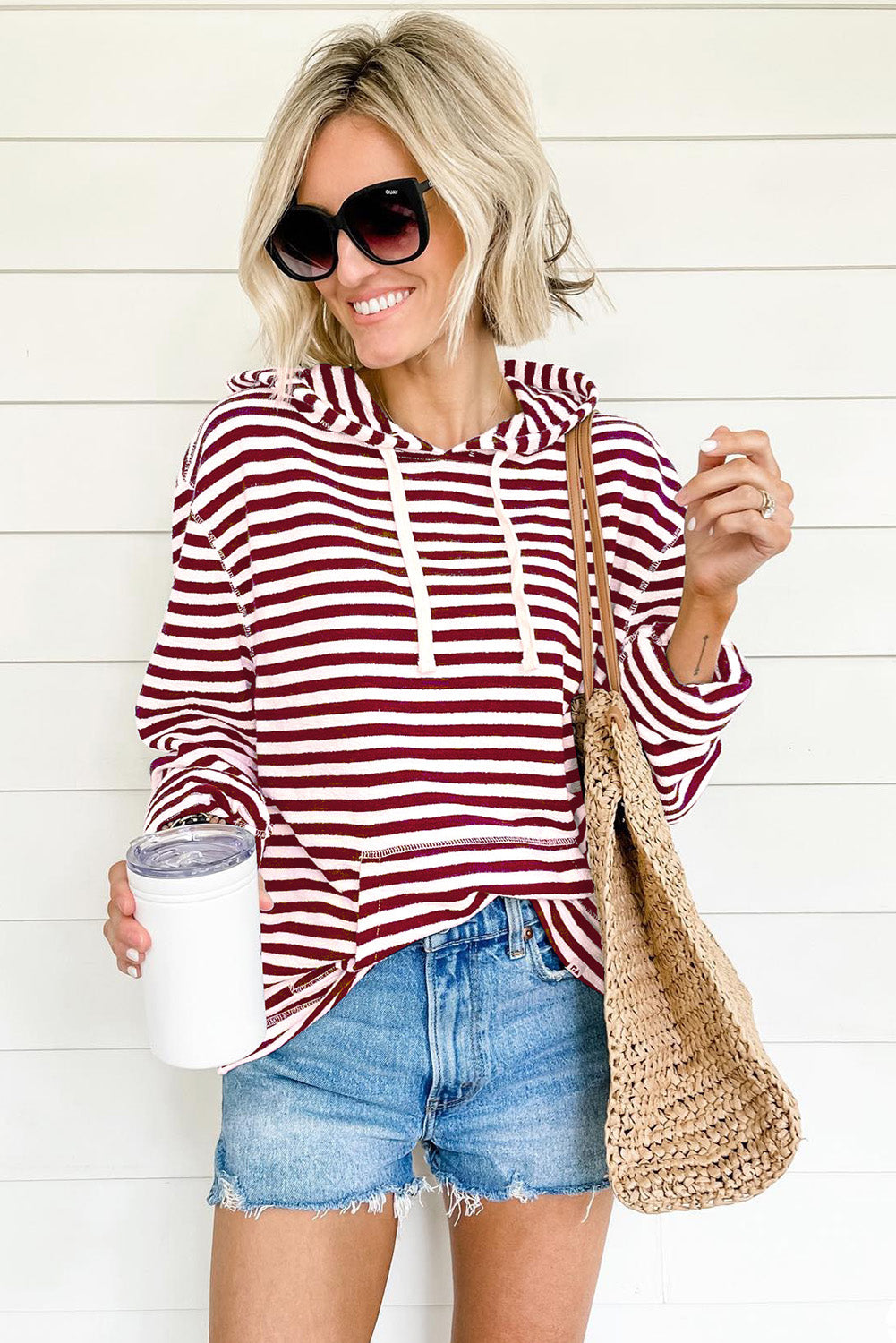 A person wearing a trendy Burgundy Stripe Kangaroo Pocket Drawstring Hoodie, paired with denim shorts and sunglasses, holds a white tumbler and a woven bag in front of a white paneled wall.