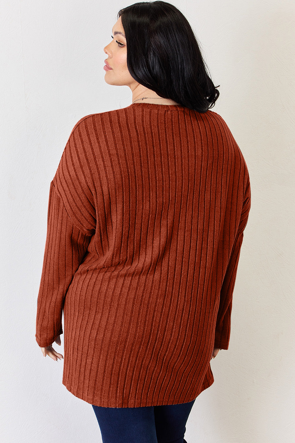 A person with long blonde hair is standing against a light background, wearing the Basic Bae Full Size Ribbed Half Button Long Sleeve High-Low T-Shirt in blue and dark pants. They are looking at the camera and smiling slightly, showcasing the chic high-low hem of their top.