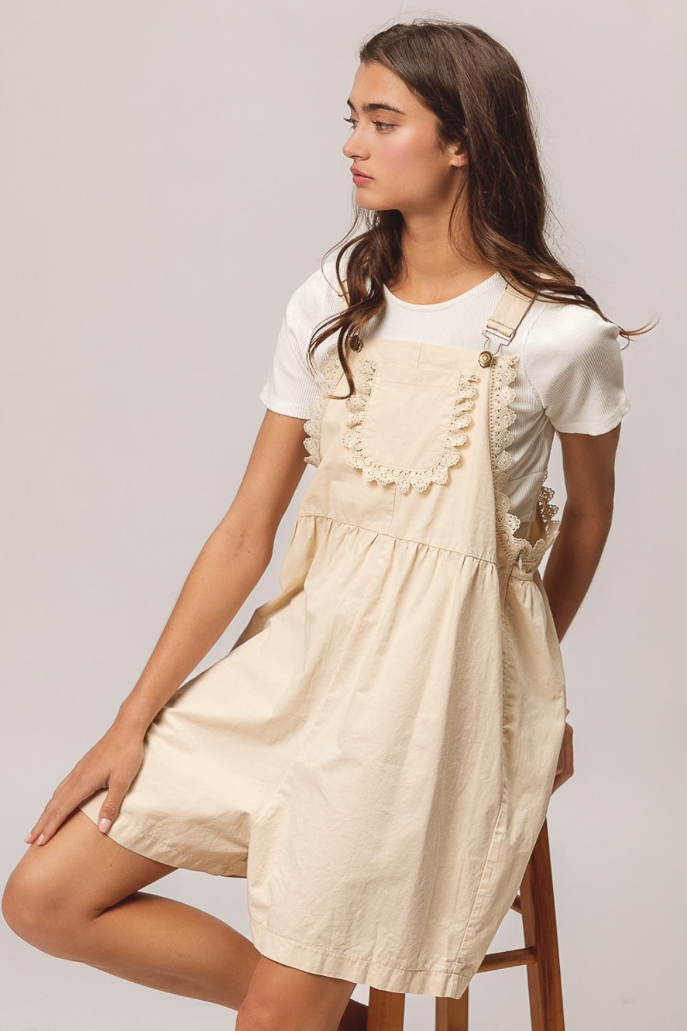 A young woman with long dark hair is sitting on a wooden stool, wearing a white t-shirt paired with the BiBi Back Patch Pocket Lace Trim Rompers—a cream-colored overall dress featuring back patch pockets. With a neutral expression set against a plain light gray background, she effortlessly embodies the perfect addition to any summer wardrobe collection.