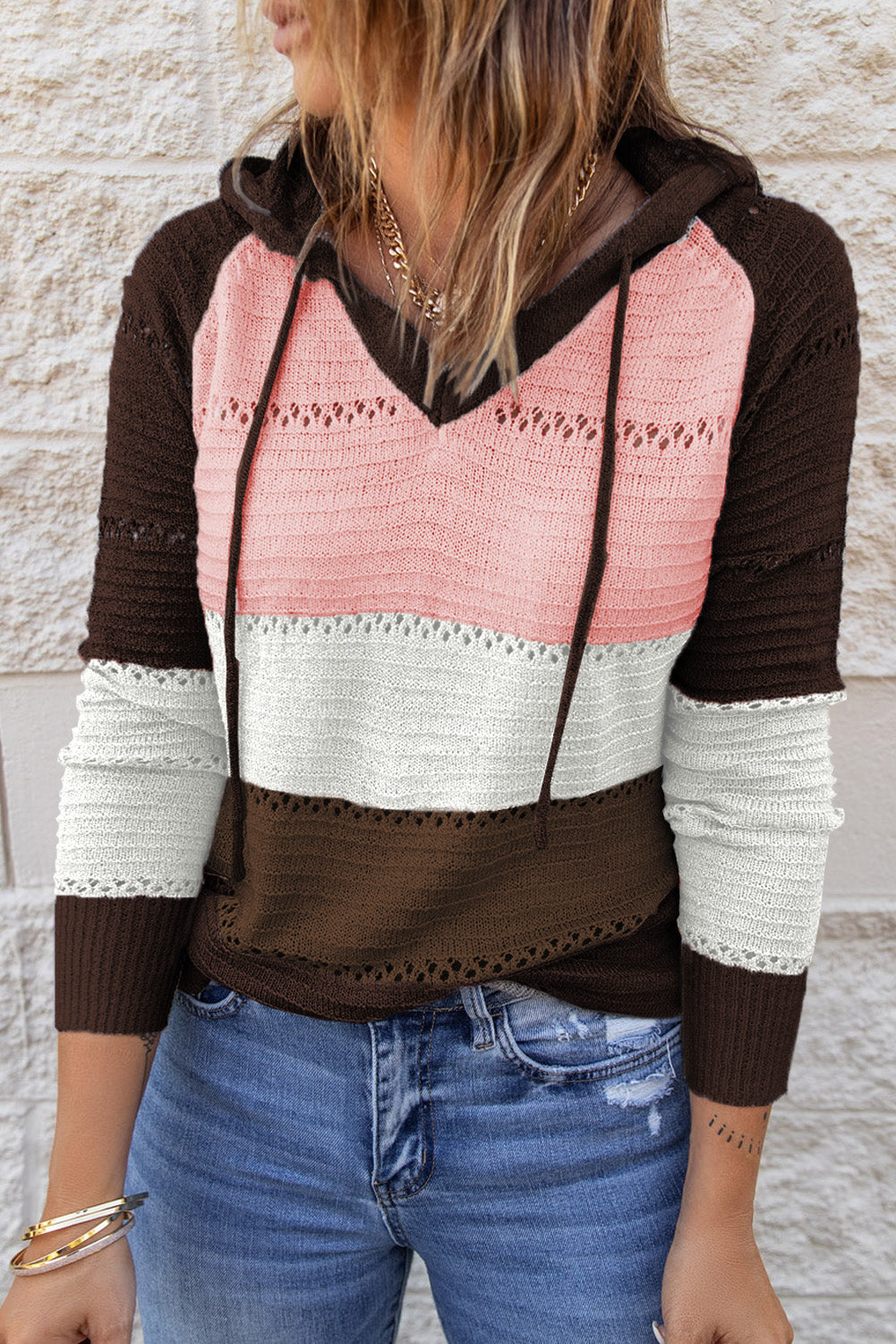 A person stands with their back to the camera, adjusting the hood of a Brown Beach Bonfire Knitted Hoodie featuring dark brown, pink, white, and light brown stripes. Perfect for a casual style, they are wearing distressed blue jeans and showcasing the laid-back vibe of lightweight hoodies.