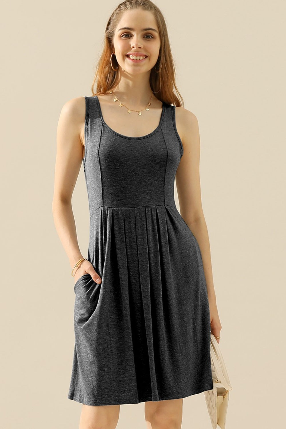 A woman in the Doublju Full Size Round Neck Ruched Sleeveless Dress with Pockets, featuring a stretchy gray fabric, smiles while holding a quilted white purse. She is wearing a gold necklace and bracelet.