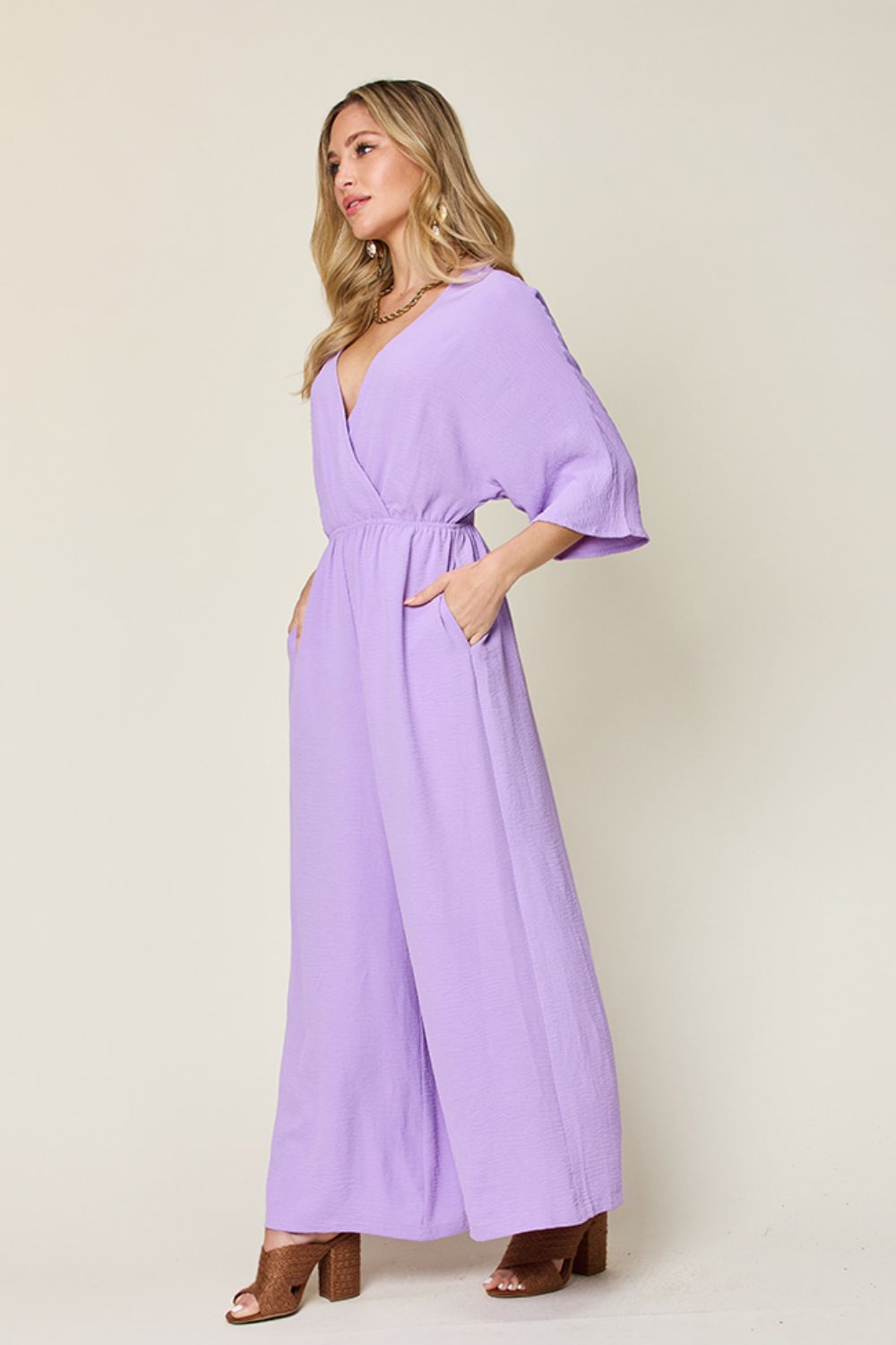 Person wearing the Double Take Full Size Surplice Wide Leg Jumpsuit with Pockets in royal blue, featuring wide legs, a deep V-neckline, and ruched detailing, posing with hands on hips against a plain light-colored background.