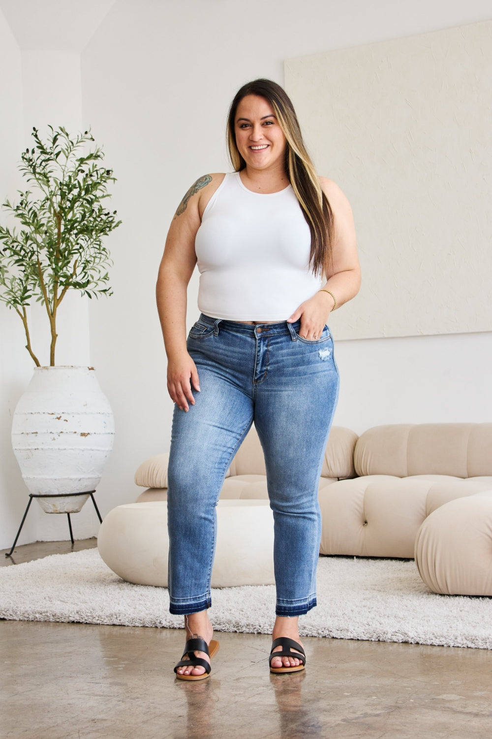 A person modeling Judy Blue Full Size Release Hem Cropped Bootcut Jeans and white heeled sandals stands in a room with light-colored furniture.