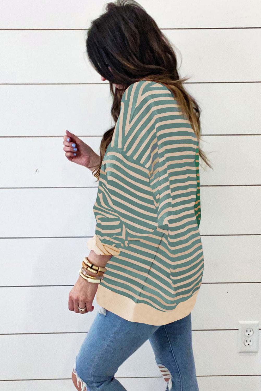 A woman with long hair is wearing a Green Stripe Casual Stripe Colorblock Drop Shoulder Oversize Sweatshirt, blue ripped jeans, and multiple bracelets, walking in front of a shiplap wall.