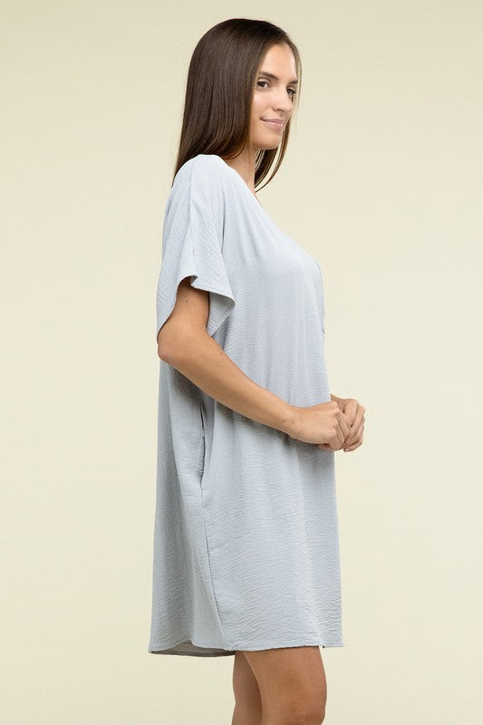 A woman wearing a loose light brown Woven Airflow V Neck T-Shirt Dress with Pockets stands against a plain background. Made from breathable fabric, this summer dress includes pockets where her hands rest as she smiles slightly.