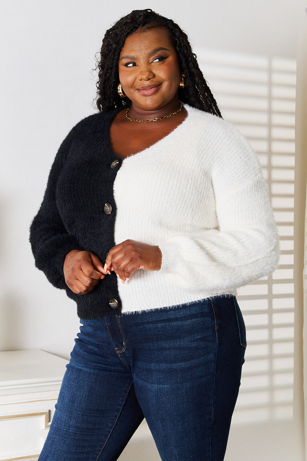 A woman stands with one hand on her hip and the other touching her face. She is wearing the Angel Wings Woven Right Contrast Button-Front V-Neck Cardigan in black and white, paired with jeans. She is smiling.