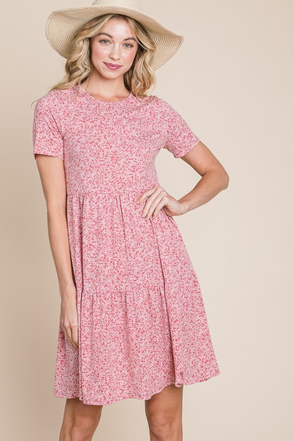 A woman with wavy blonde hair is smiling and holding the side of her BOMBOM Printed Short Sleeve Mini Dress, a lightweight summer dress featuring a pink pattern.