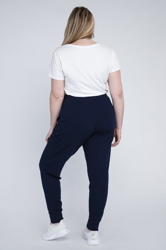 A person stands against a white background, wearing a white t-shirt, grey Plus-Size Jogger Pants with an adjustable waistband, and white sneakers. They have long blonde hair and are posing with one hand on their hip.