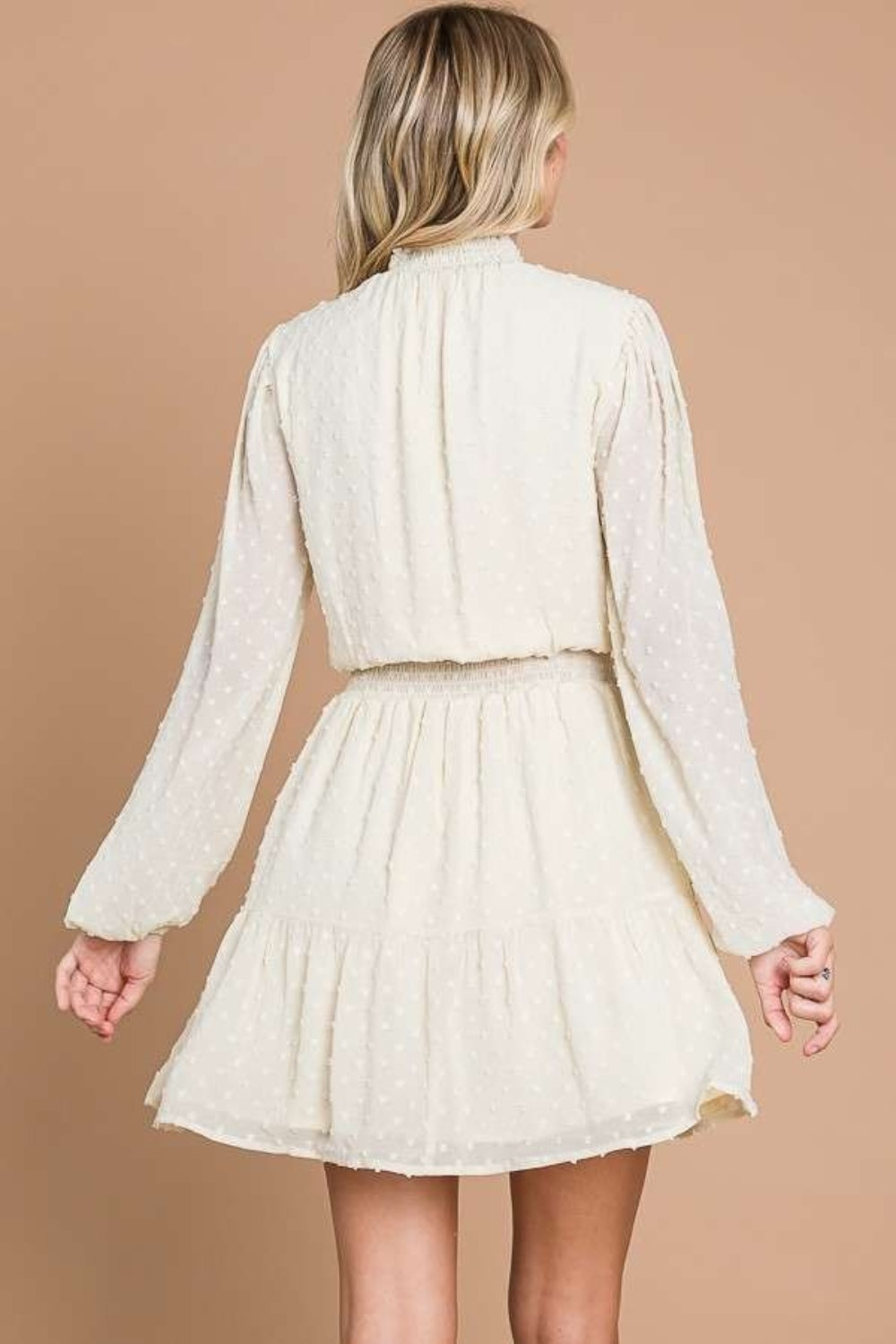 Wearing the Culture Code Swiss Dot Smock Neck Mini Dress with Pockets in light beige, a person stands against a brown backdrop, smiling, with one hand on the hip and the other gently holding the elegantly draped lightweight chiffon fabric.