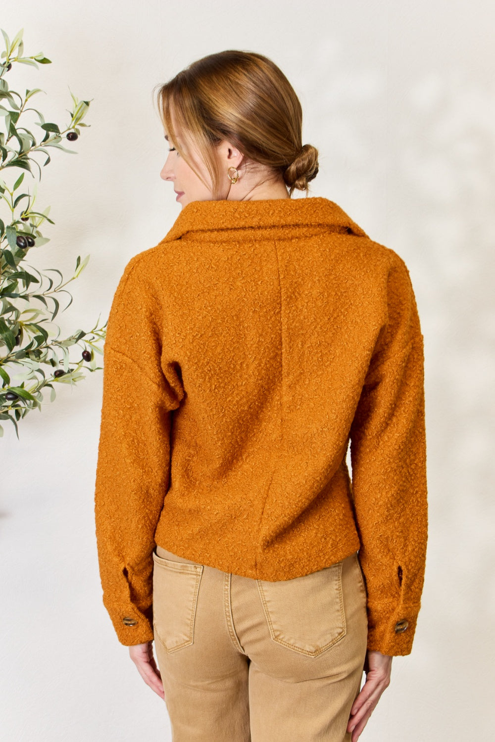 A person wears a brown textured Culture Code Full Size Half Button Turtleneck Sweatshirt made from breathable fabrics and tan pants, standing next to a leafy plant against a light background.