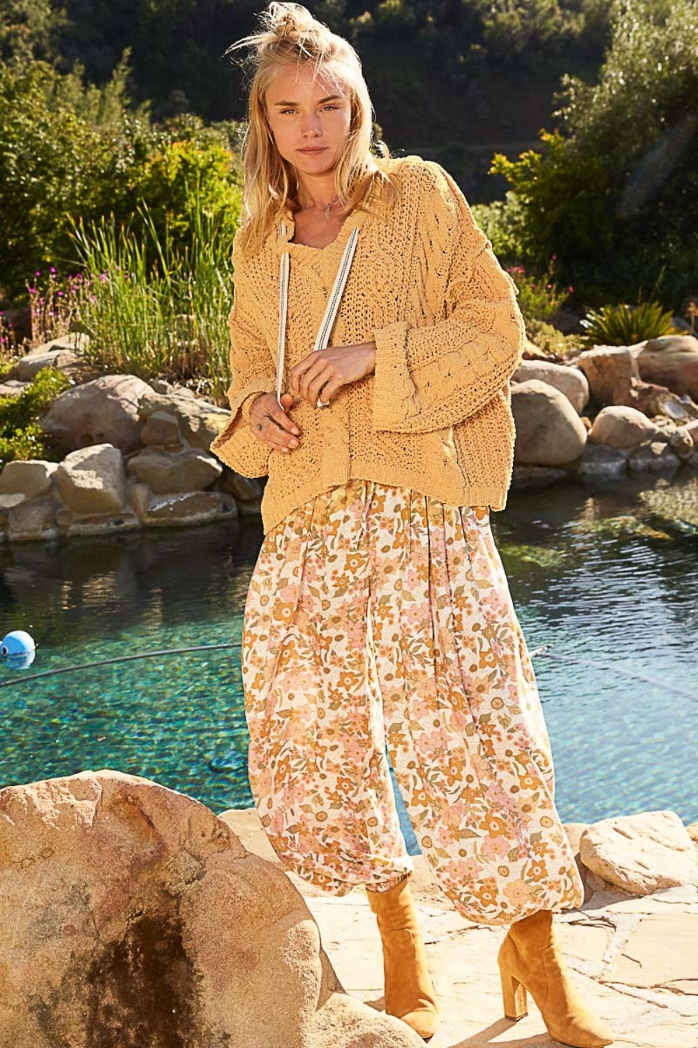 A person with long blonde hair, wearing a cozy and stylish POL Cable Knit Hooded Chenille Sweater paired with a patterned skirt, smiles outdoors with greenery in the background.