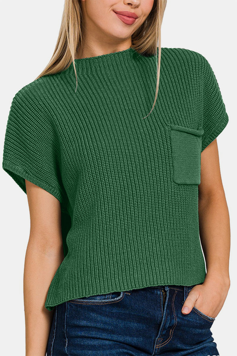 A woman wearing the Zenana Mock Neck Short Sleeve Cropped Sweater in green, featuring a small front pocket and crafted from knit fabric, is standing and smiling. She has long, straight hair and is paired with dark blue jeans.