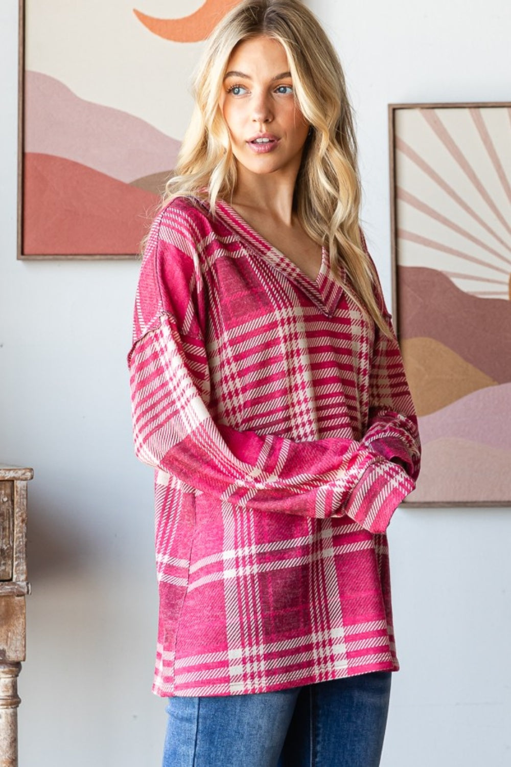 A person with long blond hair smiles while wearing the Heimish Plaid Drop Shoulder V-Neck T-Shirt in red and white and a pair of blue jeans, leaning against a wooden table with framed pictures in the background.