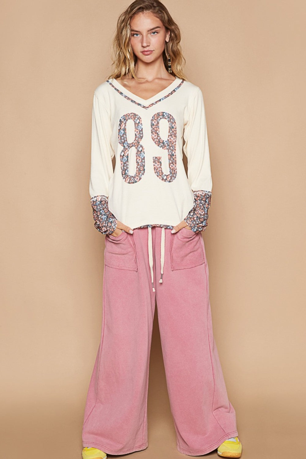 A young woman with blonde hair wears a POL V-Neck Long Sleeve Floral Number Patch T-Shirt, adorned with the number 89 in a floral design on the front and matching floral patterns on the cuffs. She also wears pink pants.