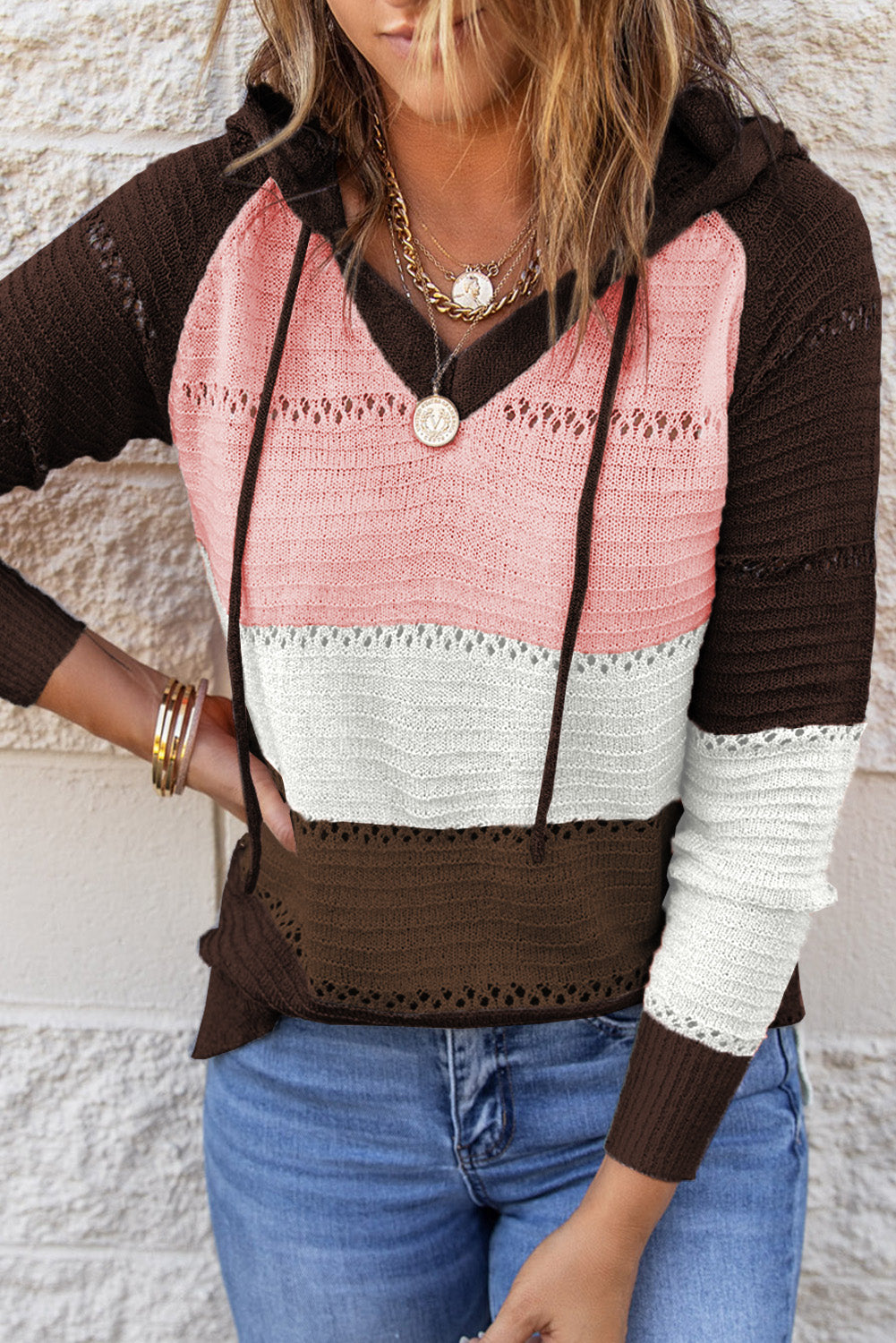 A person stands with their back to the camera, adjusting the hood of a Brown Beach Bonfire Knitted Hoodie featuring dark brown, pink, white, and light brown stripes. Perfect for a casual style, they are wearing distressed blue jeans and showcasing the laid-back vibe of lightweight hoodies.