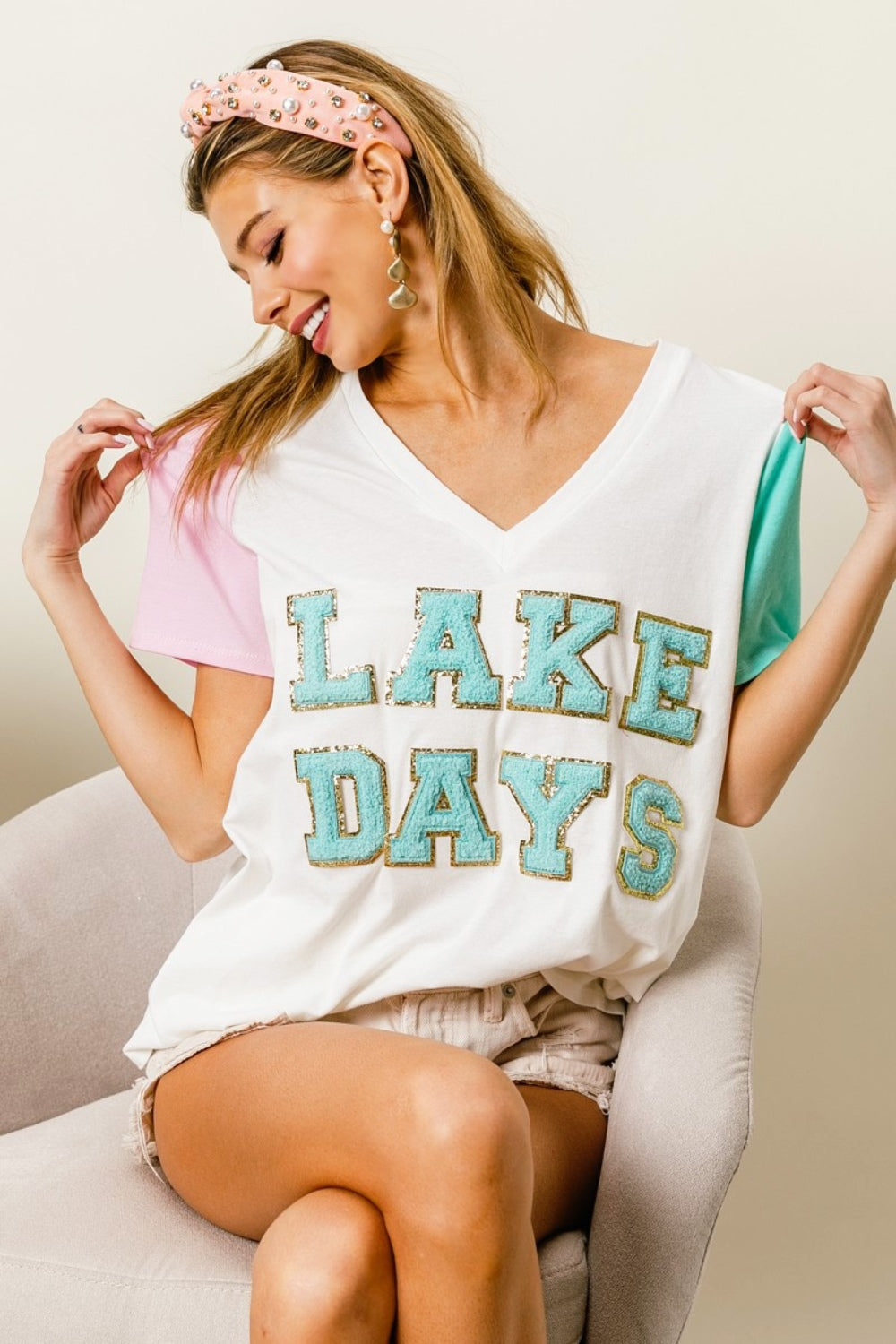 A woman wearing a BiBi Letter Patch V-Neck Color Block Short Sleeve T-Shirt, which features "Lake Days" printed on it, sits on a chair. She holds the shirt's sleeves and wears a headband and earrings.