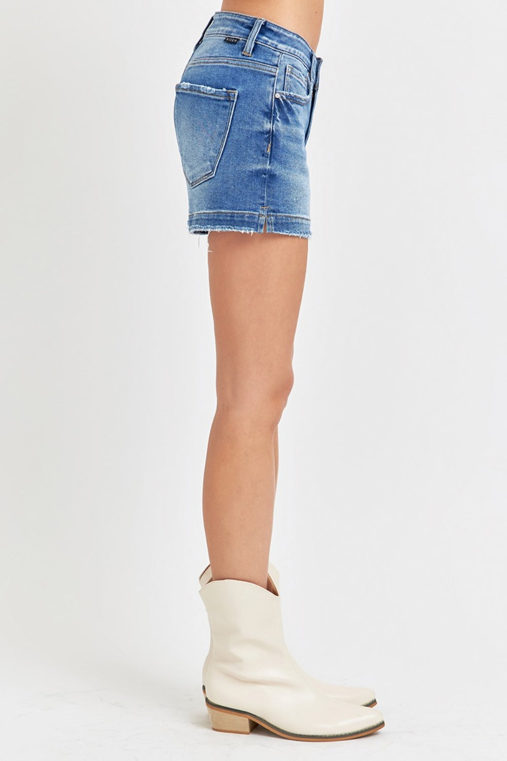 A person wearing RISEN Low Rise Slit Denim Shorts with hands in pockets, accessorized with a white bracelet.