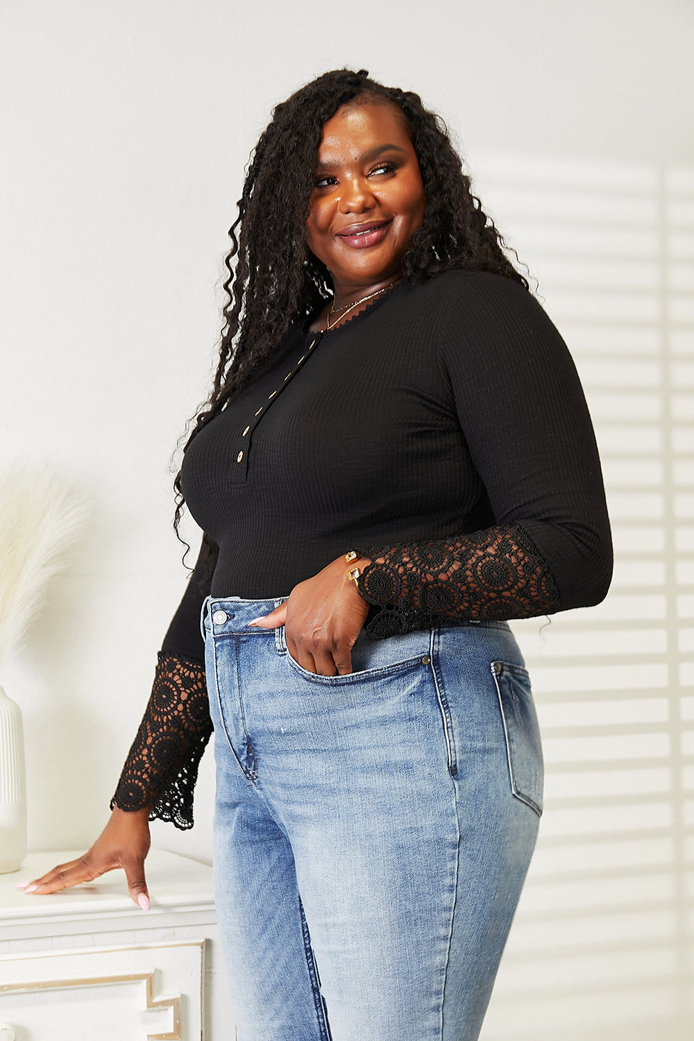 A woman with long curly hair wears the Double Take Crochet Lace Hem Sleeve Button Top and versatile solid color blue jeans, standing indoors with a neutral expression.