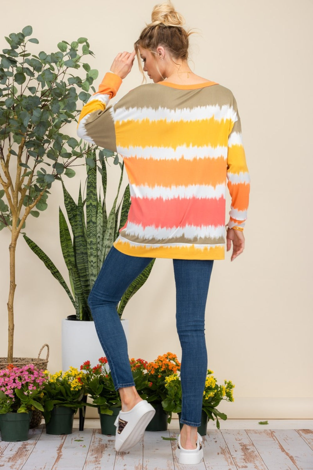 Wearing the Celeste Full Size Striped Long Sleeve T-Shirt paired with jeans, a person smiles while standing against a light background adorned with plants, exuding a casual yet stylish look.