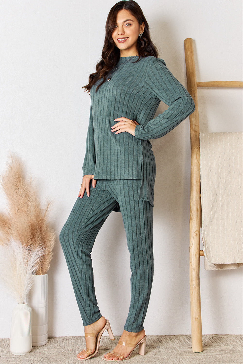 A woman in a Basic Bae Full Size Ribbed Round Neck High-Low Slit Top and Pants Set poses indoors near a wooden ladder. She is wearing the matching maroon slightly stretchy top and pants, paired with beige heels. Pampas grass in a white vase is visible in the foreground. For care, machine wash cold.