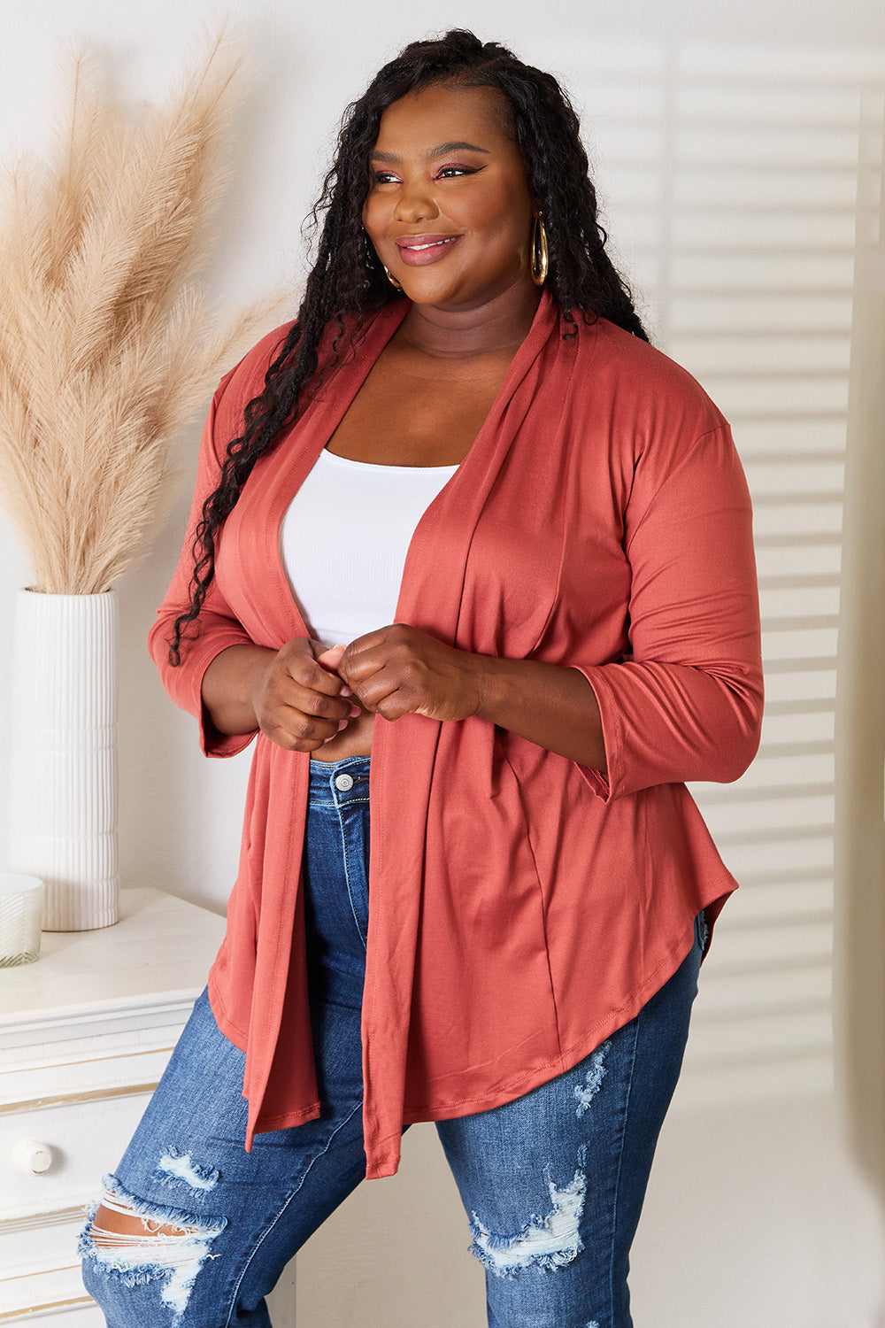 A person with long hair is wearing the Culture Code Full Size Open Front Cardigan in a rust color, complemented by a flattering draped front. They pair it with a white top and ripped jeans while standing indoors with one hand on their hip.