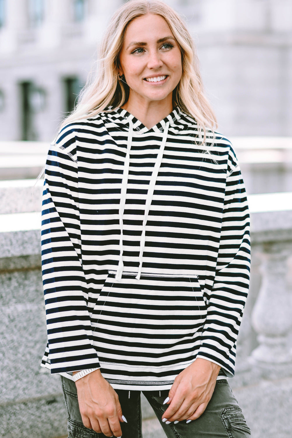 A person with long, blonde hair wearing a trendy Stripe Kangaroo Pocket Drawstring Hoodie with black and white stripes and dark green pants is standing outdoors, facing away from the camera.