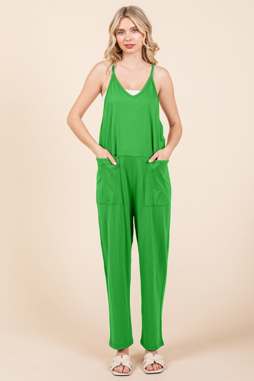 A person stands against a plain background wearing the Culture Code Full Size Sleeveless Jumpsuit with Pockets in green, paired with white sandals, showcasing its practical pockets with hands tucked comfortably inside.