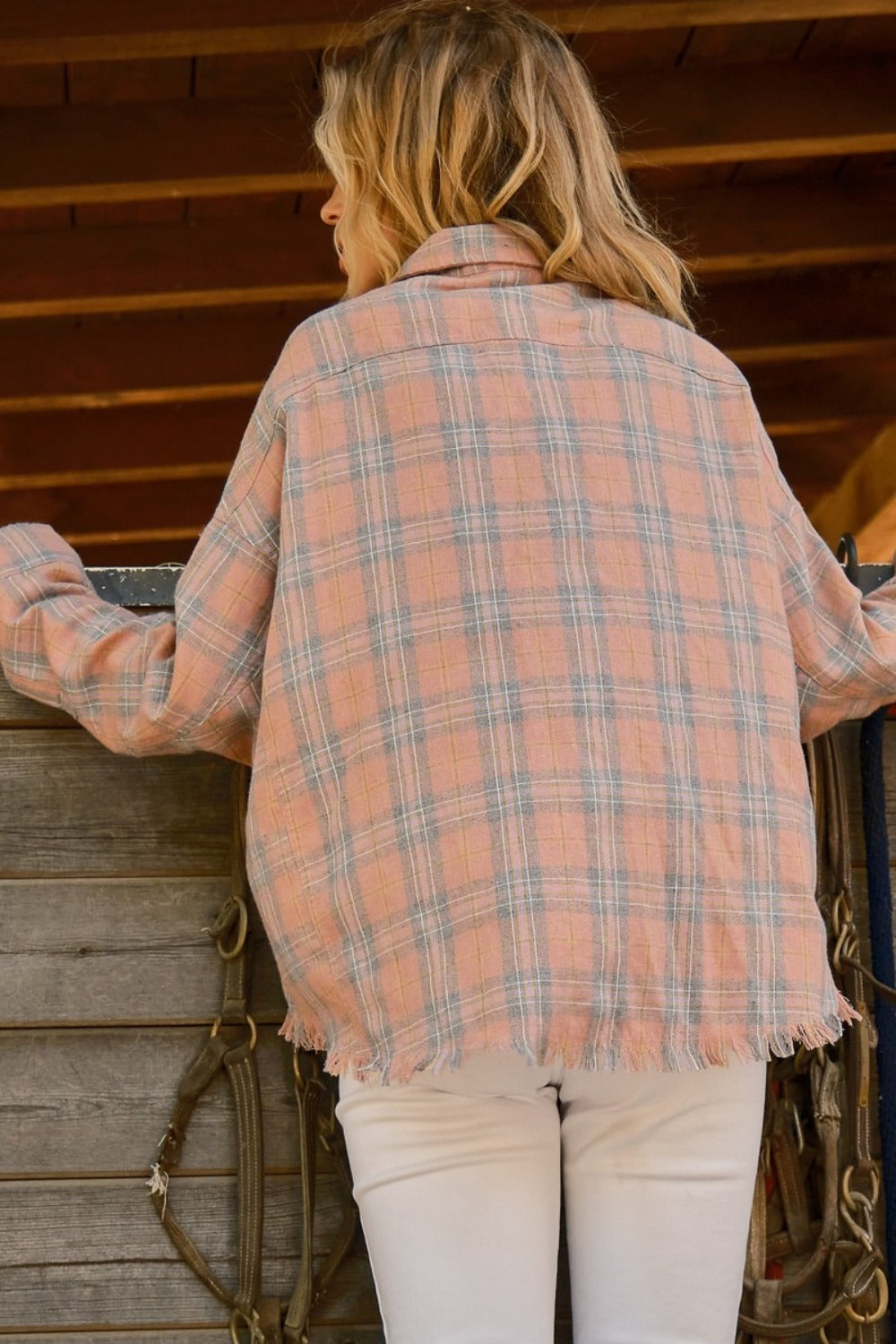 A person wearing the And The Why Full Size Plaid Button Up Raw Hem Shirt and white pants is standing in front of a wooden structure, showcasing raw edge detailing, with bridles hanging beside them.