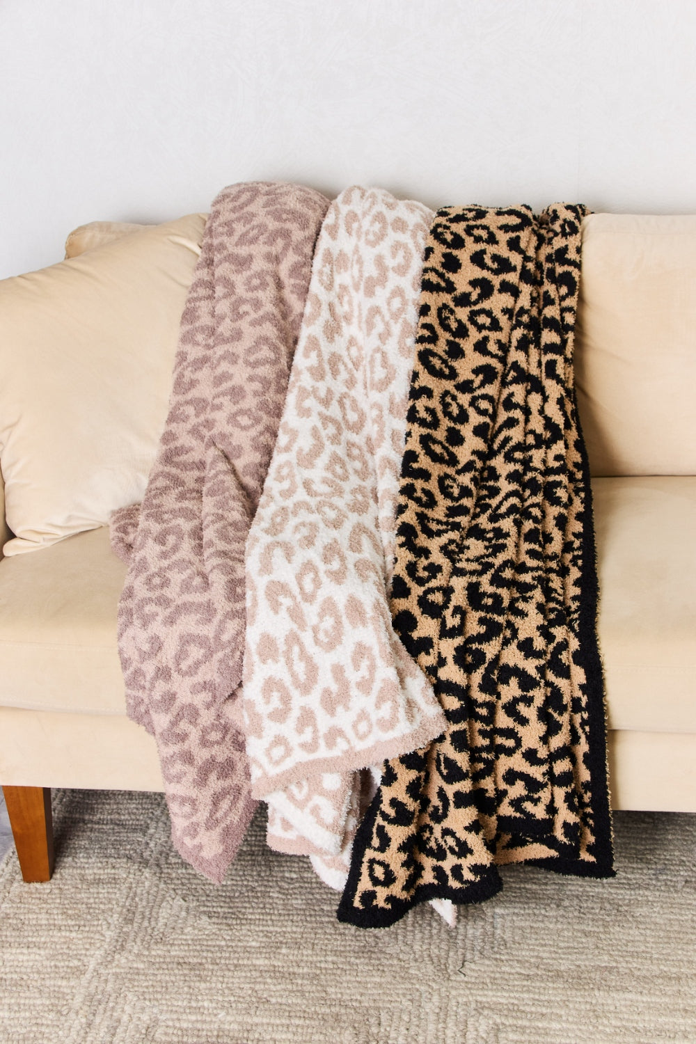 A woman stands in a room holding up the Cuddley Leopard Decorative Throw Blanket, which features a large, leopard-print design in a neutral color scheme. She is smiling and facing the camera, showcasing the 100% polyester fabric.