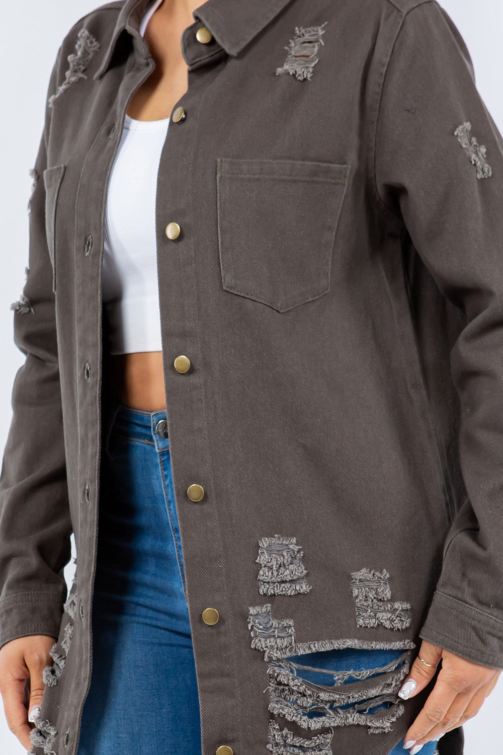 A woman stands against a light background wearing the American Bazi Distressed Button Up Long Sleeve Denim Jacket with a white crop top and blue ripped jeans.