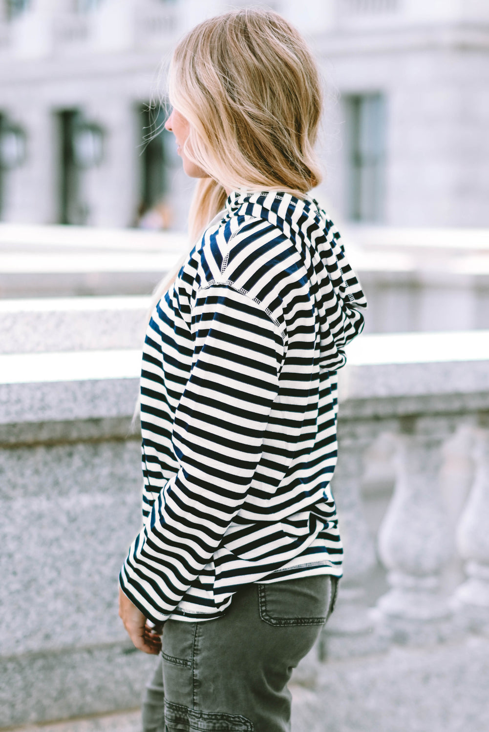 A person with long, blonde hair wearing a trendy Stripe Kangaroo Pocket Drawstring Hoodie with black and white stripes and dark green pants is standing outdoors, facing away from the camera.