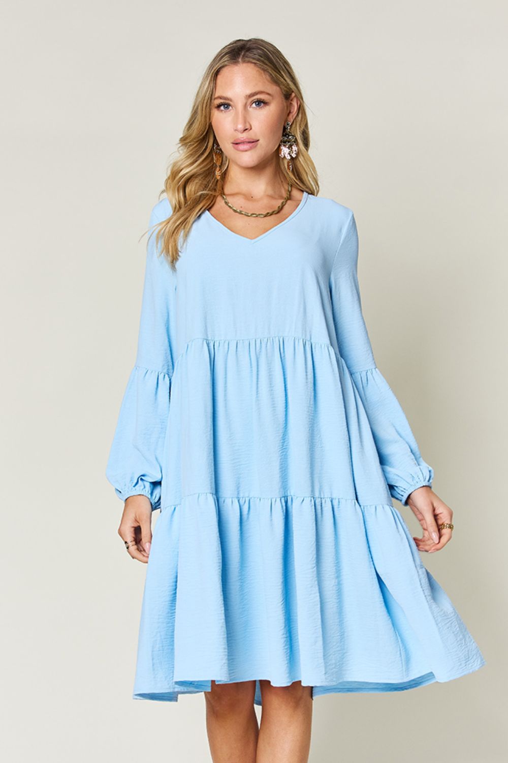 A woman stands wearing the Double Take Full Size V-Neck Balloon Sleeve Tiered Dress with Pockets in pink. Crafted from high stretch yarn, this dress combines comfort and style effortlessly. She has long, wavy hair and accessorizes with hoop earrings and a necklace. This imported garment is easy to care for – simply machine wash cold to keep it looking fresh.