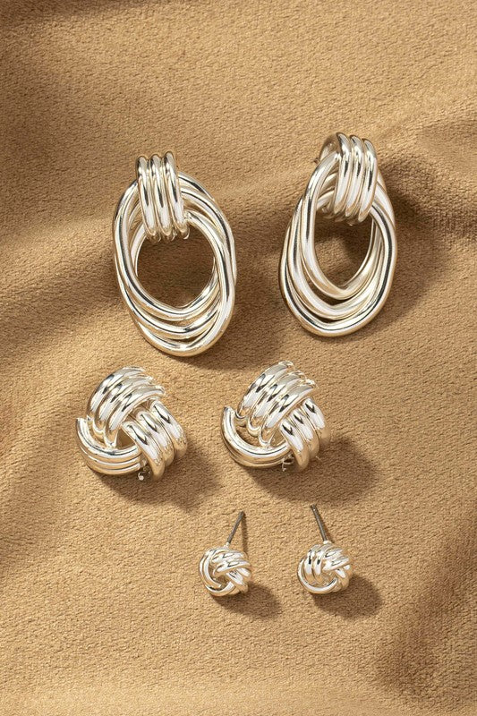 Close-up of a person wearing the large, shiny gold-plated Premium Trio Metal Knot and Hoop Earrings with their finger touching it. Their skin and hair are visible, and they are wearing a white shirt, making these earrings ideal for both casual and formal occasions.