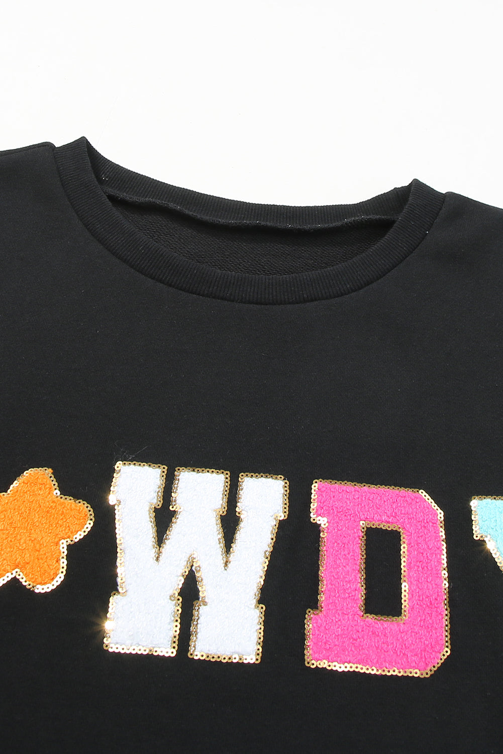 A woman with short blonde hair is wearing the Black Howdy Glitter Chenille Patch Graphic Casual Sweatshirt, featuring colorful star patterns and "WD" letters. She pairs it with light denim shorts and smiles while looking to the side.